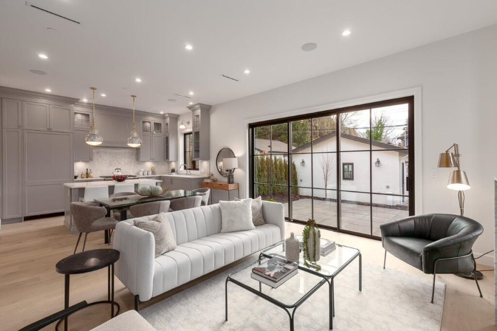 A bright living room with large picture windows revealing the backyard. A grey modern renovated kitchen behind the sofa in an open concept living space.