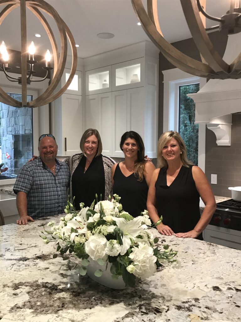 Happy clients and a happy designer stand around a stone kitchen island with a white flower centrepiece.