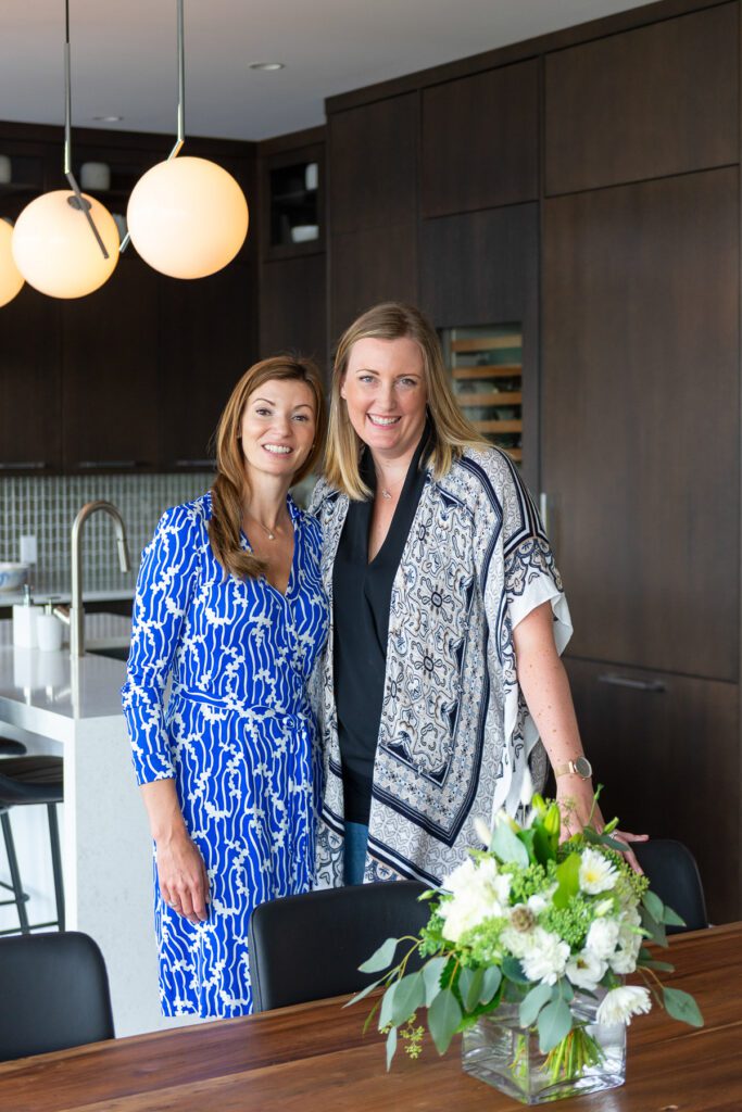 Sarah and a happy client stand in a renovated dining room with spherical drop lighting and a dark hardwood table.