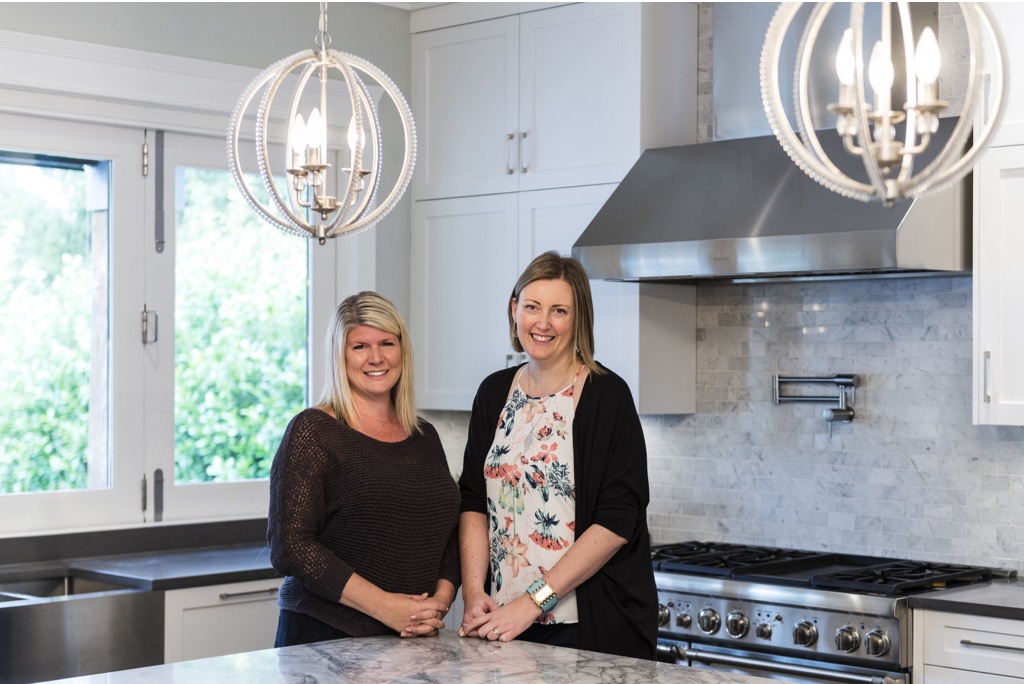 Sarah and a happy client stand in a freshly renovated kitchen with round drop lighting and stainless hood fan.