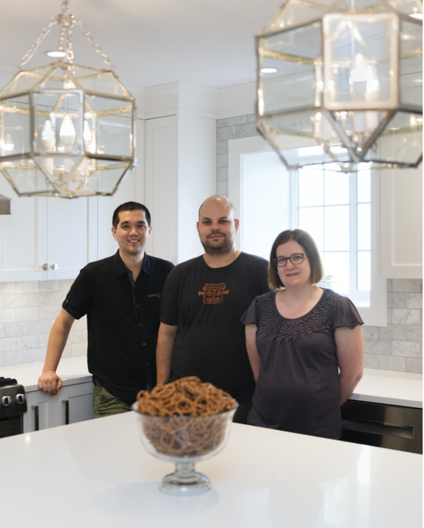 Three people stand in a beautifully renovated kitchen behind the island under glass chandeliers.