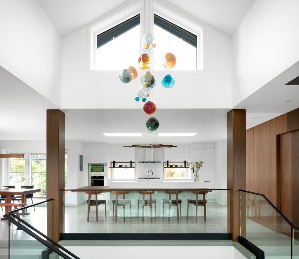 A modern renovated kitchen with unique colourful chandelier over a kitchen island with seating looking into the bright kitchen framed with wooden support columns.