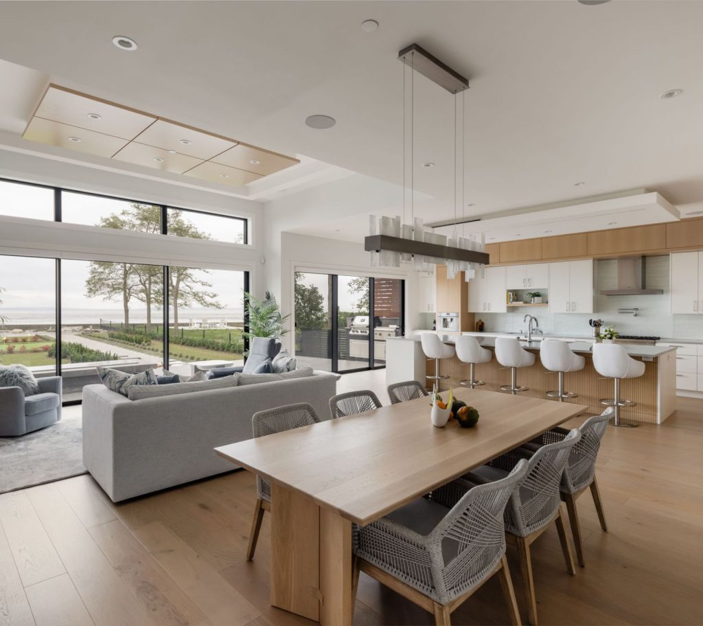An open concept space including a dining area for six in the foreground, a kitchen with barstools in the background, and a cozy grey couch facing out to the picture windows looking out the expansive view.